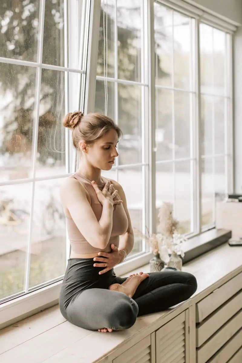 A Woman in Beige Tank Top Sitting Near the Glass Windows while Meditating