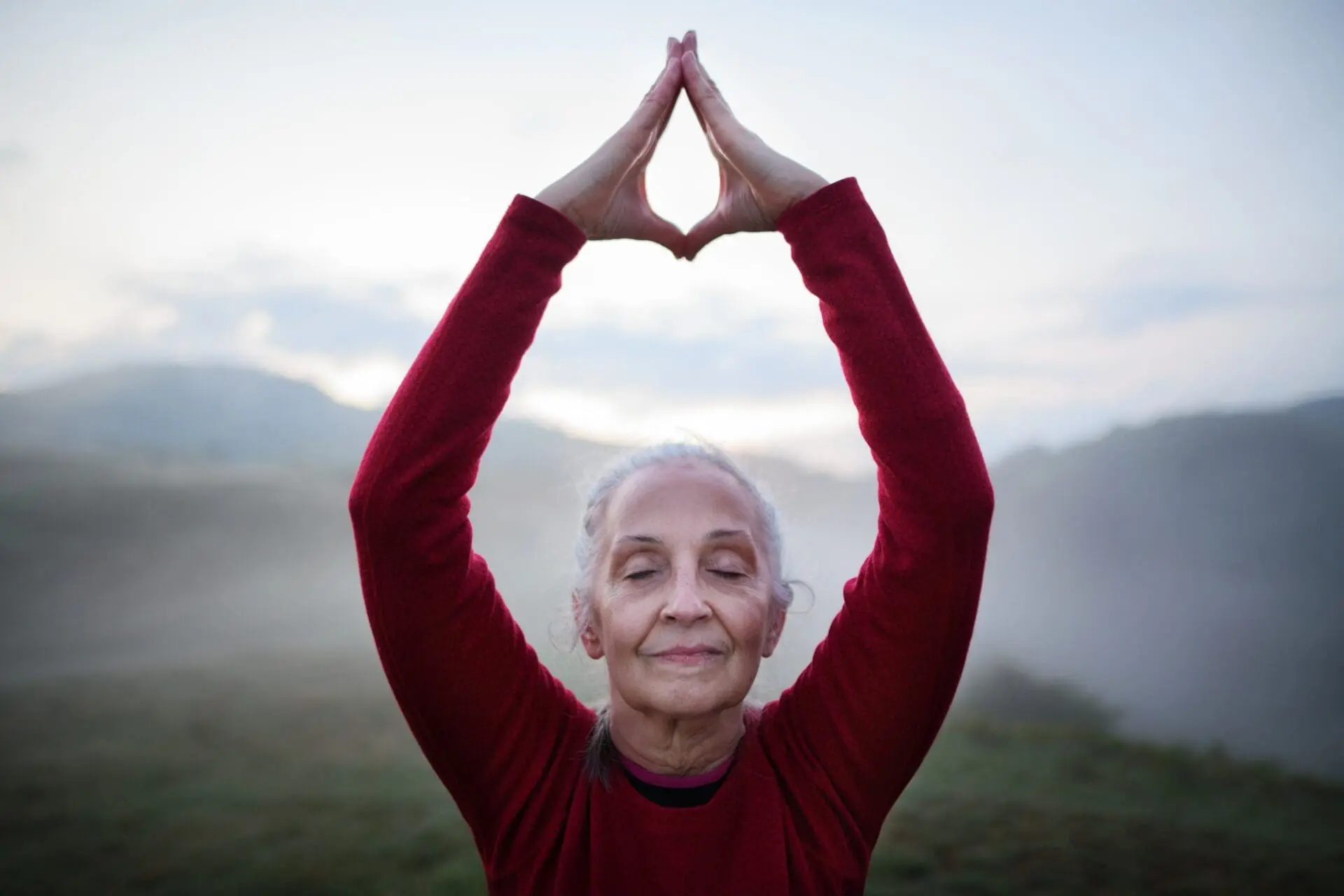 Senior woman doing breathing exercise in nature on early morning with fog and mountains in
